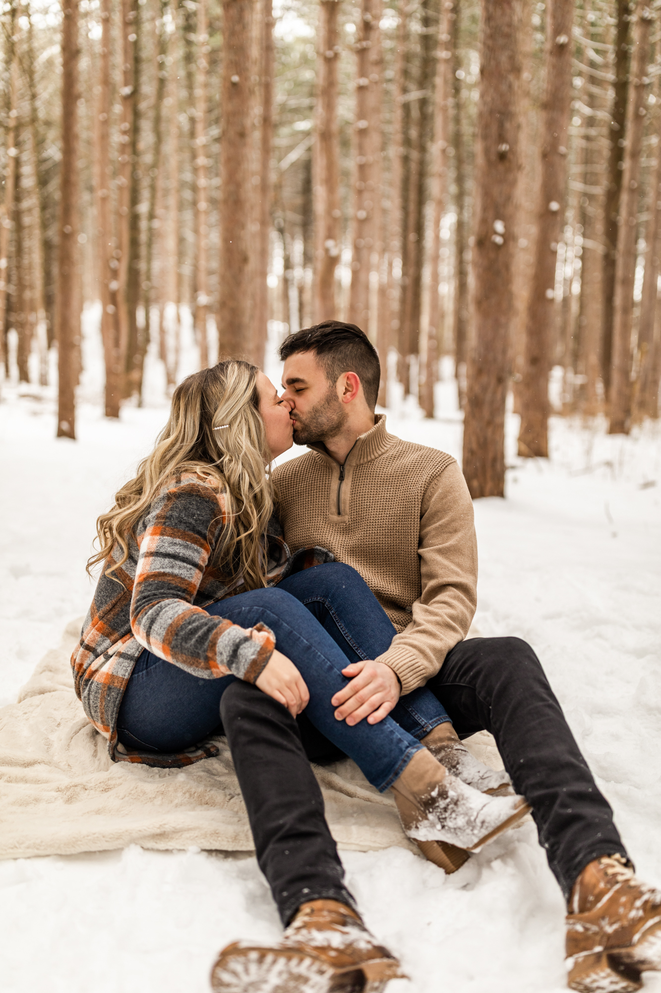 Couple sitting on blanket and kissing in snowy woods for winter engagement photos in Michigan