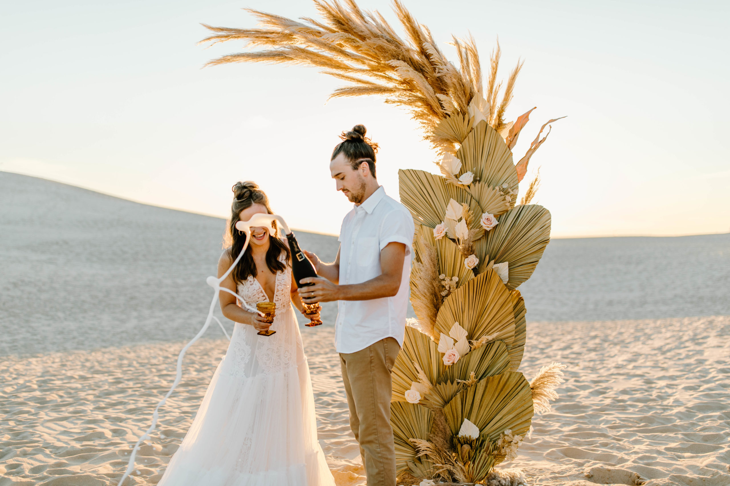 couple popping champagne at Sand Dune Elopement in front of Palms and Pampas Grass