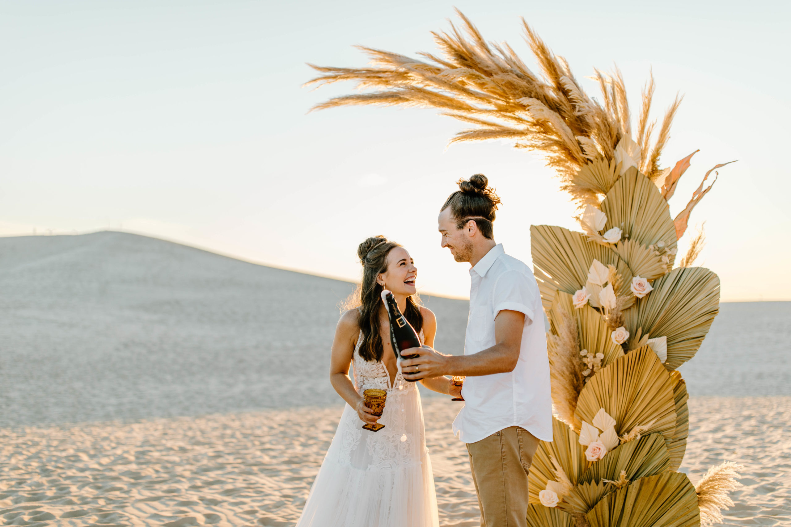 couple popping champagne at Sand Dune Elopement in front of Palms and Pampas Grass