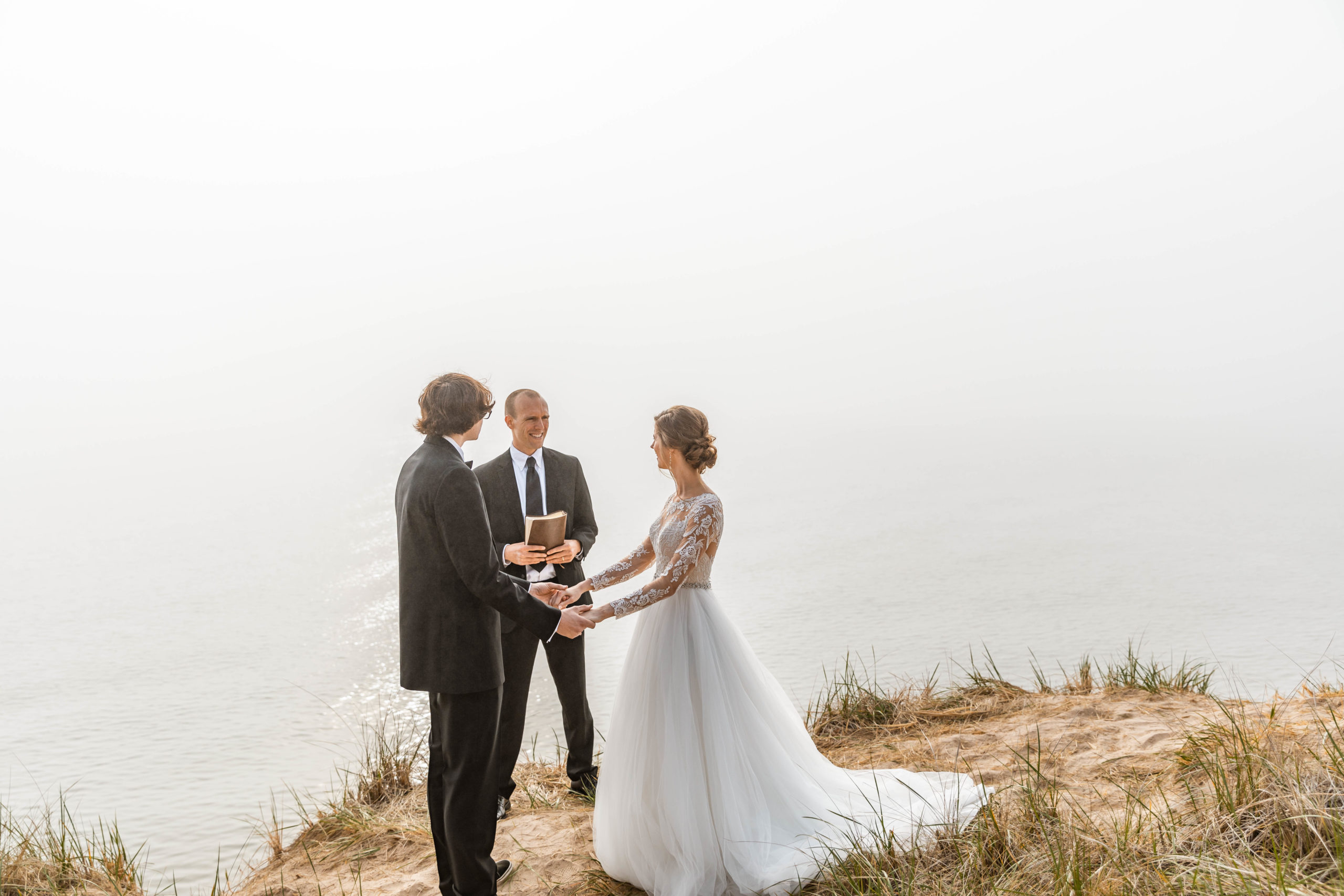 Bride and groom holding hands in front of officiant on dunes overlooking lake