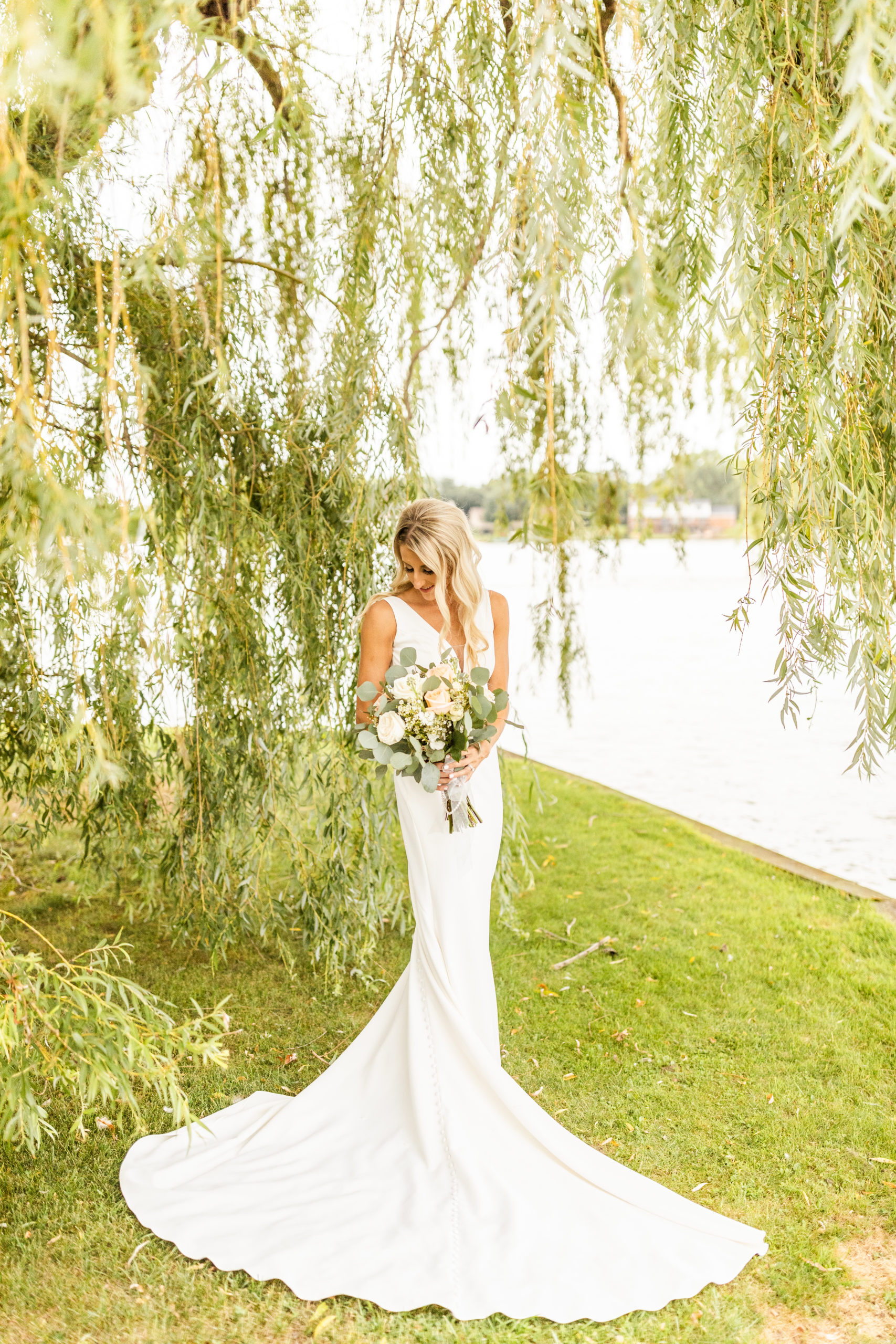 bride posing under lakefront willow tree holding bouquet in Michigan wedding