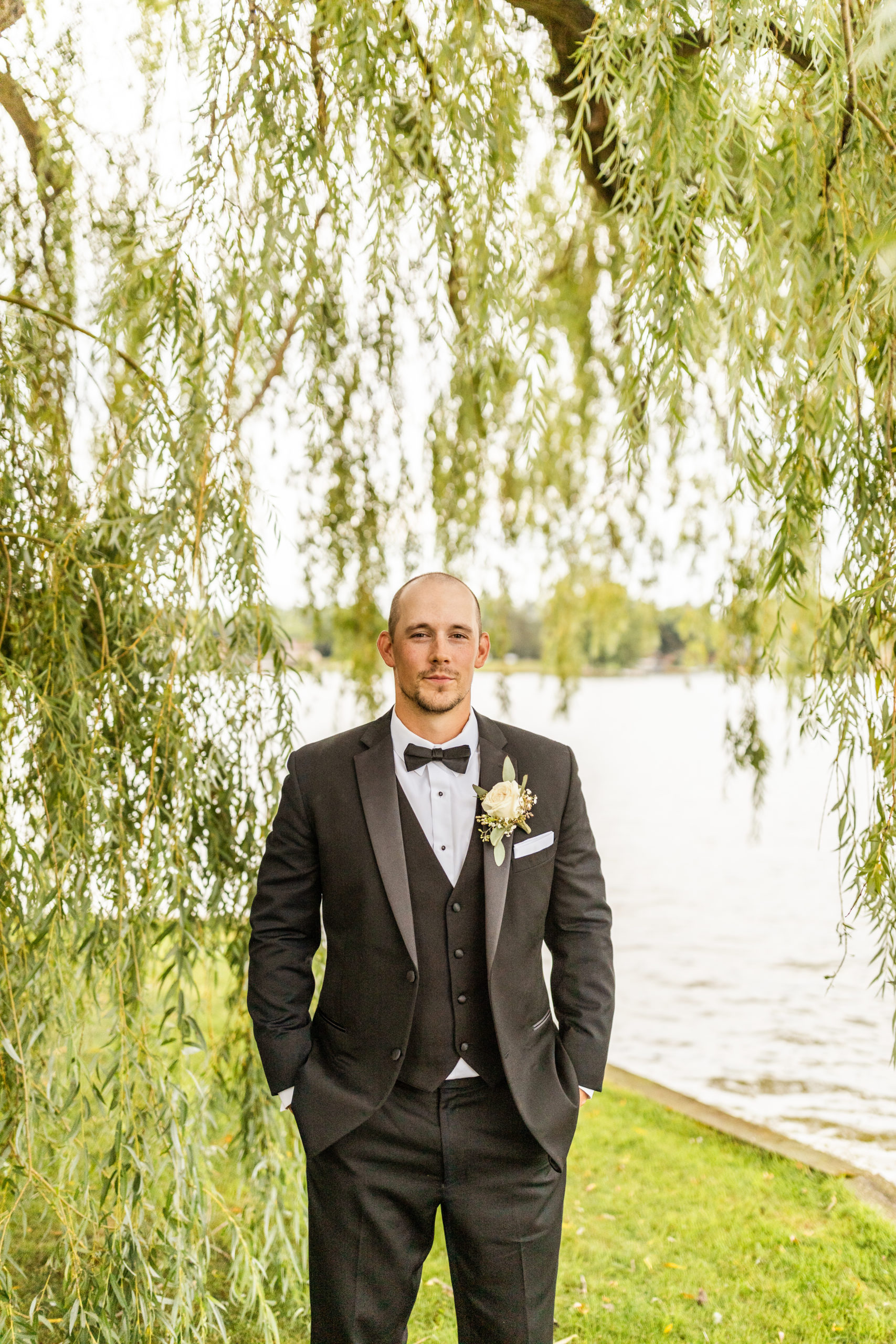 groom standing under lakefront willow tree in black tuxedo in wedding photos